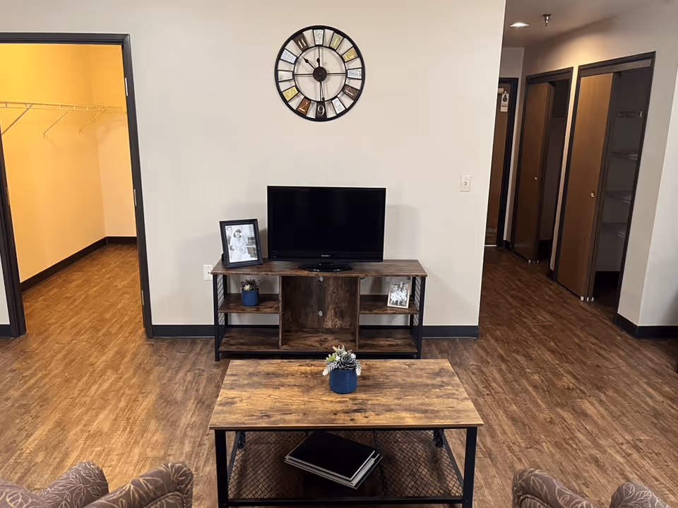 Interior view of a living room area with wooden flooring, a wooden TV stand holding a flat-screen TV and framed photos, a wooden coffee table with a small plant, and a large wall clock above the TV. There are open doorways leading to other rooms, including a walk-in closet on the left and additional rooms with wooden doors on the right.