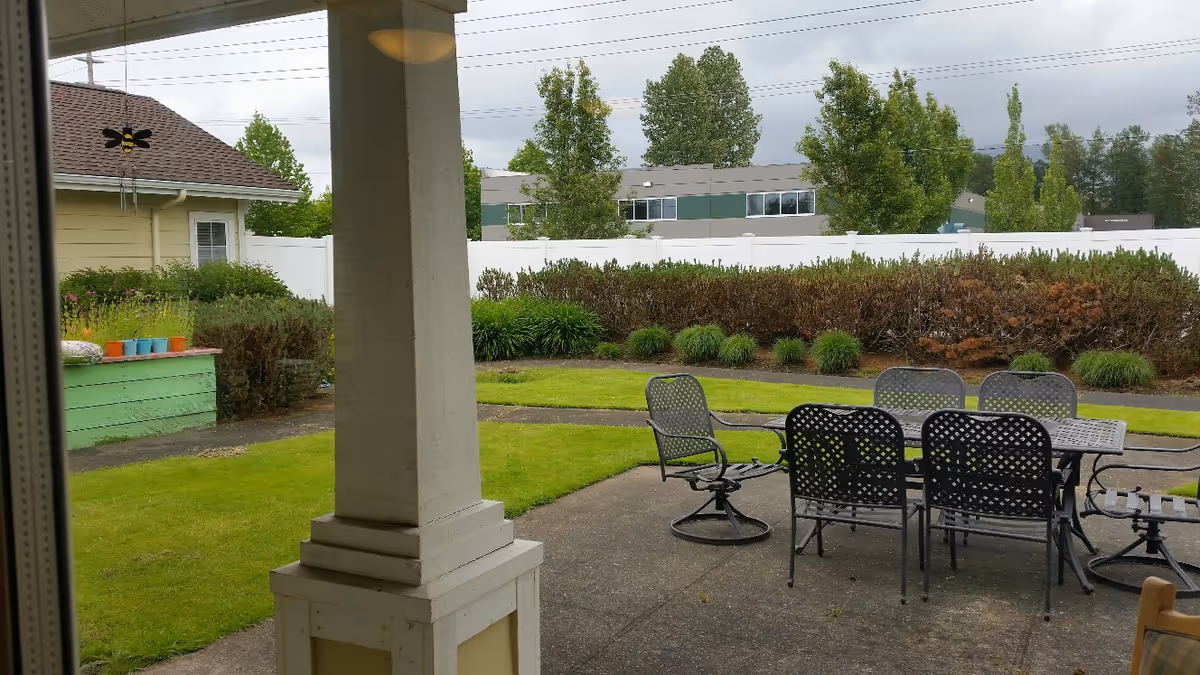 Covered patio with metal dining table and chairs overlooking a grassy yard, planter boxes, and neighboring buildings.