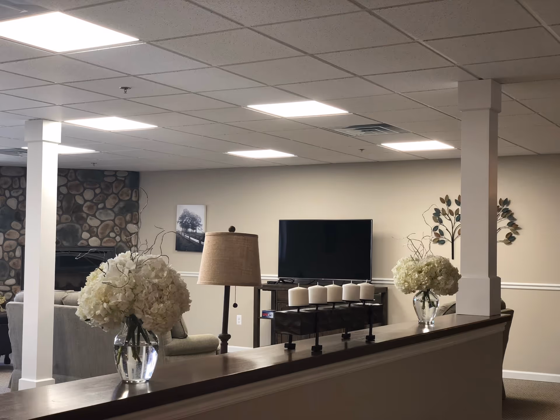 A cozy senior living room with beige walls and carpeted floor. The room features a stone fireplace, a flat-screen TV on a wooden stand, two vases with white flowers on a half wall, a table lamp, and decorative wall art including a black and white photo and a metal tree sculpture.