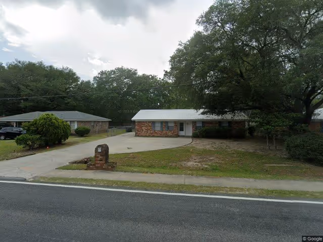 Single-story brick residential building with a white roof, a concrete driveway, and a small brick mailbox in front. The building is surrounded by trees and bushes, with a paved road in the foreground.