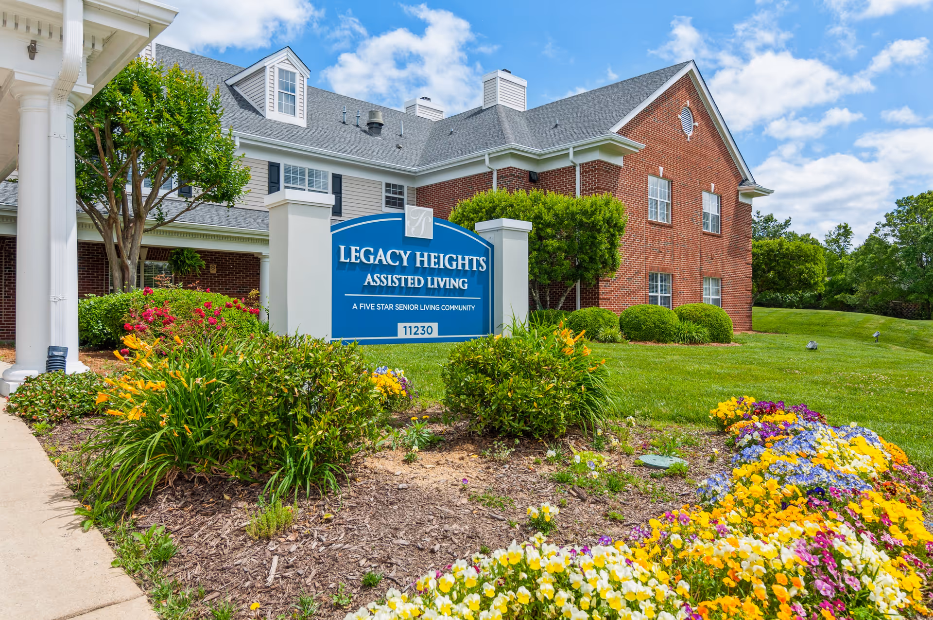 Exterior view of Legacy Heights Assisted Living community building with a blue sign in front surrounded by colorful flowers and green shrubs under a partly cloudy sky.