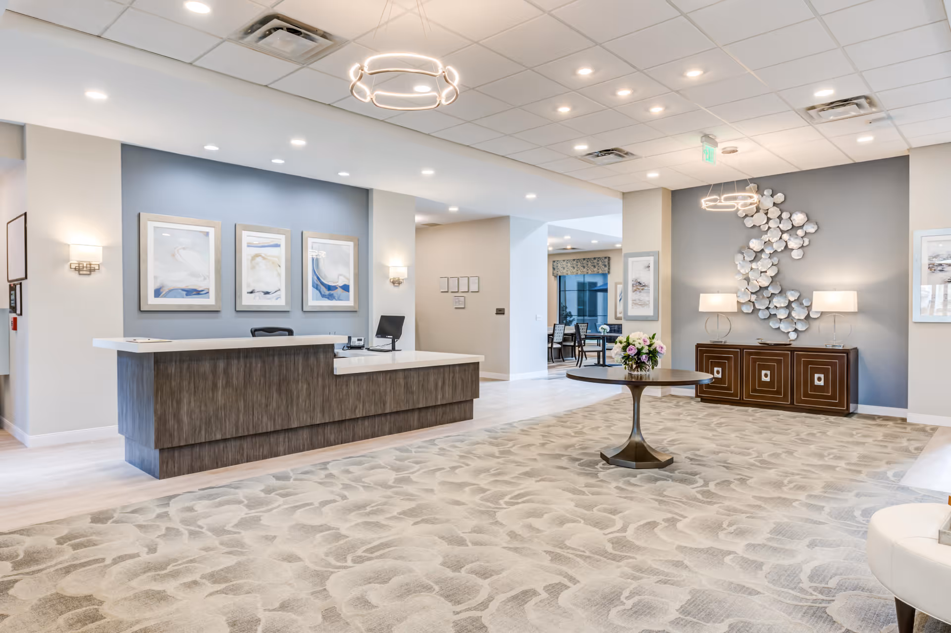 A spacious and modern reception area with a large wooden front desk, three framed artworks on the wall behind it, and a round table with a flower arrangement in the center of a patterned carpet. The room features soft lighting, decorative wall art, and a sideboard with two lamps against a gray accent wall.