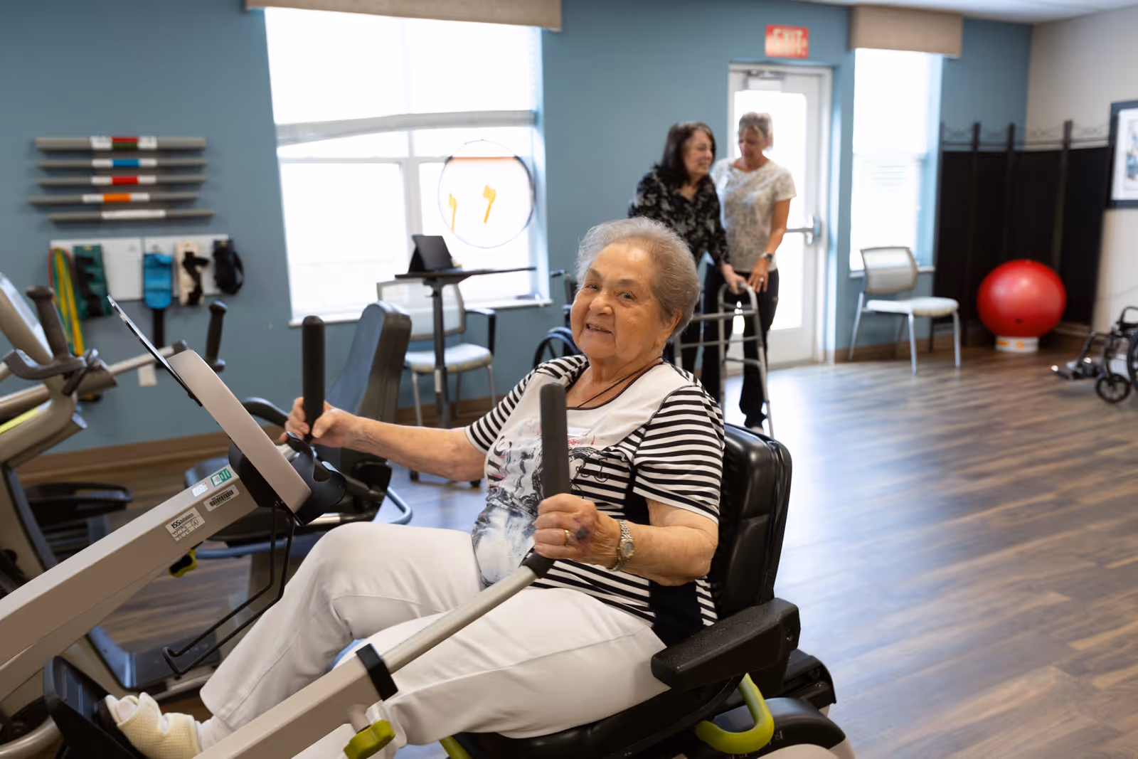 An elderly woman using a seated exercise machine in a fitness room with two women in the background, one assisting the other with a walker. The room has wooden flooring, exercise equipment, chairs, and a large red exercise ball near the wall.