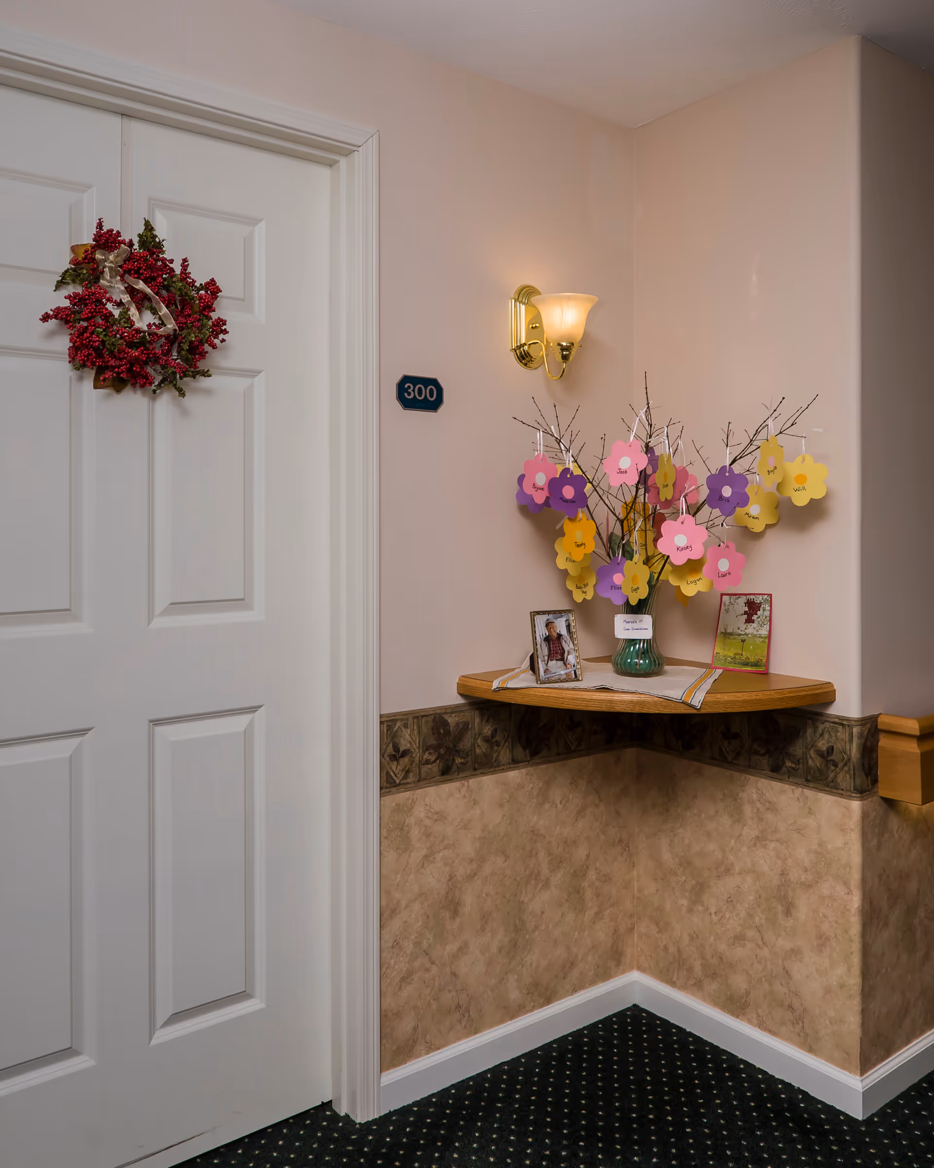 Interior corner of a hallway in a senior living facility with a white door decorated with a red berry wreath, a wall-mounted light fixture, and a small wooden corner shelf holding a vase with branches adorned with colorful paper flowers and two framed photos.