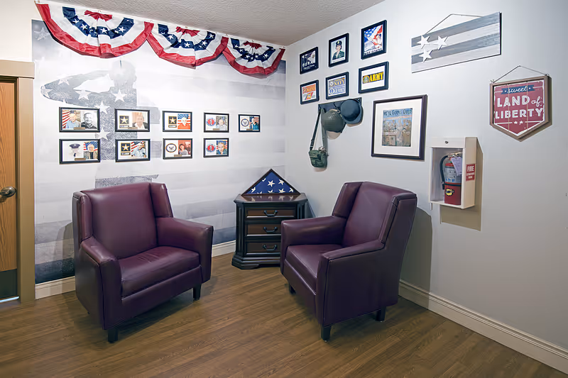 Two burgundy armchairs in a small patriotic-themed sitting area with framed photos, a folded American flag on a side table, and bunting on the wall.