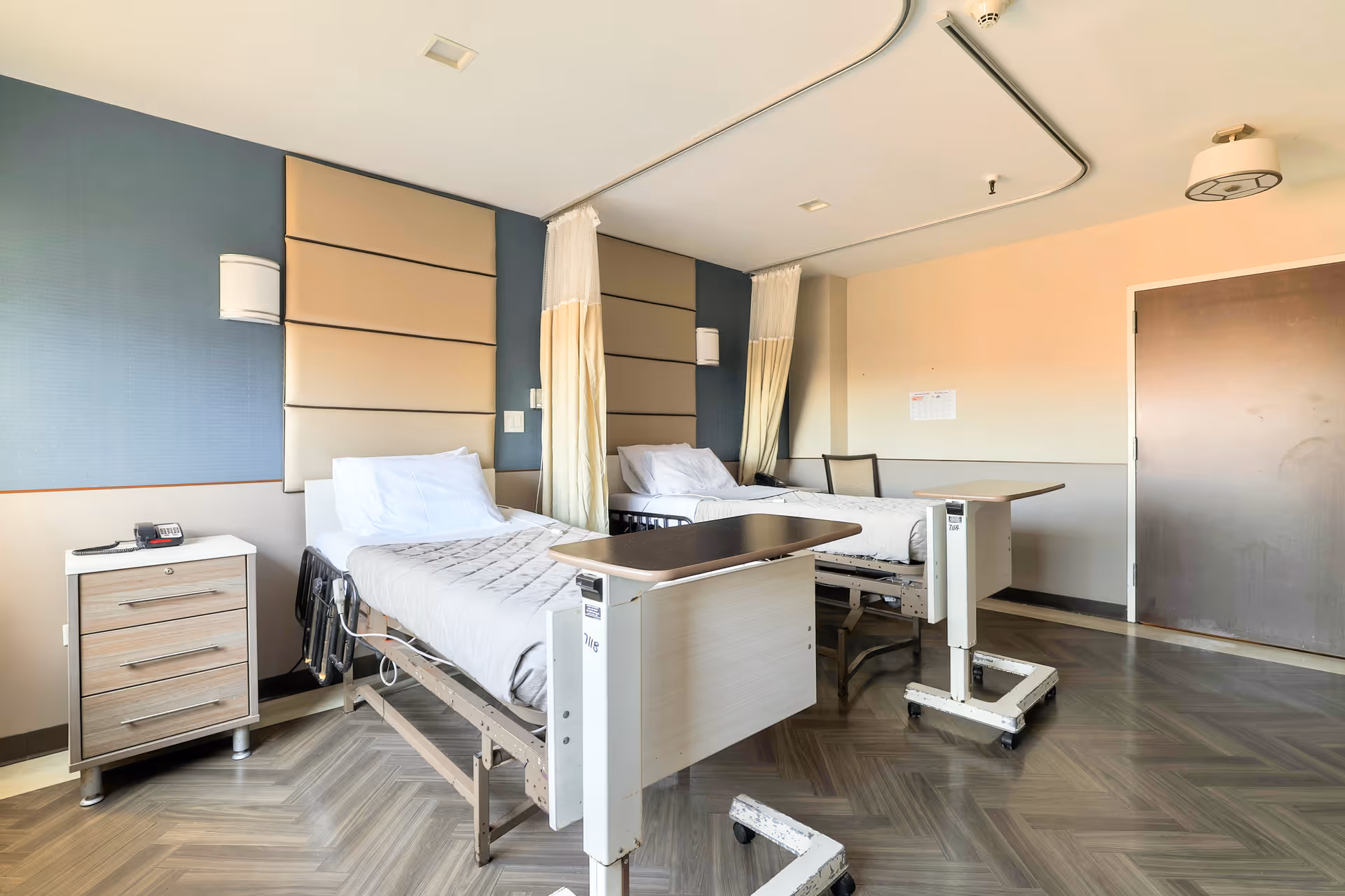 A bright and clean nursing facility room with two hospital beds, each equipped with white pillows and beige blankets. Each bed has an adjustable overbed table and a privacy curtain. There is a small wooden nightstand with drawers and a phone on it next to one of the beds. The walls are painted in neutral tones with padded headboards behind the beds, and the floor has a herringbone pattern.