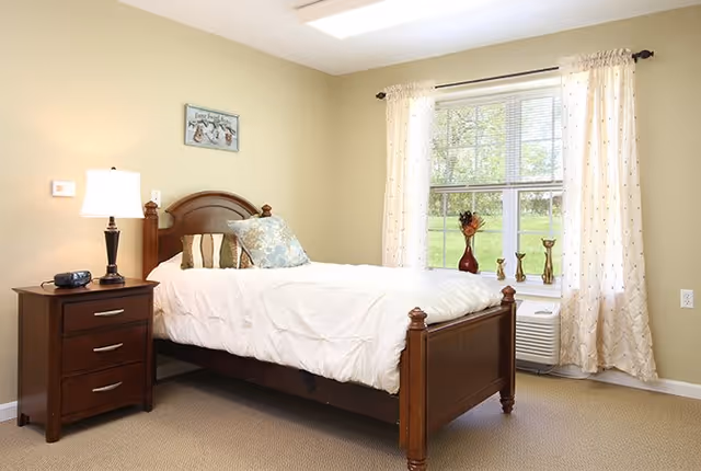 A bright bedroom with a wooden bed frame and white bedding. There are decorative pillows on the bed and a wooden nightstand with a lamp and a clock radio. A window with sheer curtains lets in natural light, and decorative items are placed on the windowsill.