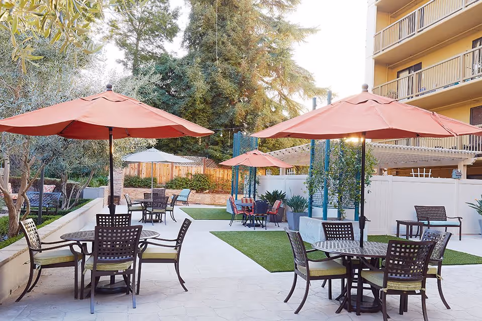Outdoor patio area with multiple round tables and chairs, each shaded by large orange umbrellas. The space is surrounded by greenery including trees and plants, with a multi-story building visible on the right side.