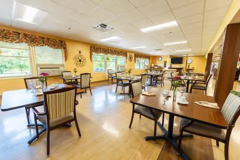 A bright dining room with multiple wooden tables and chairs arranged neatly. Each table is set with glasses, napkins, and small flower vases. Large windows with decorative valances allow natural light to fill the room. The walls are painted yellow, and there is a television mounted on the far wall.