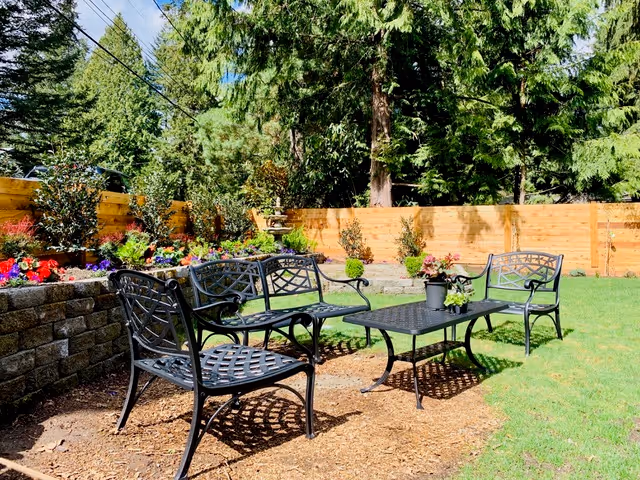 Outdoor garden area with black metal chairs and a matching table on a mulch ground, surrounded by green grass, colorful flowers, a wooden fence, and tall trees in the background under a clear sky.