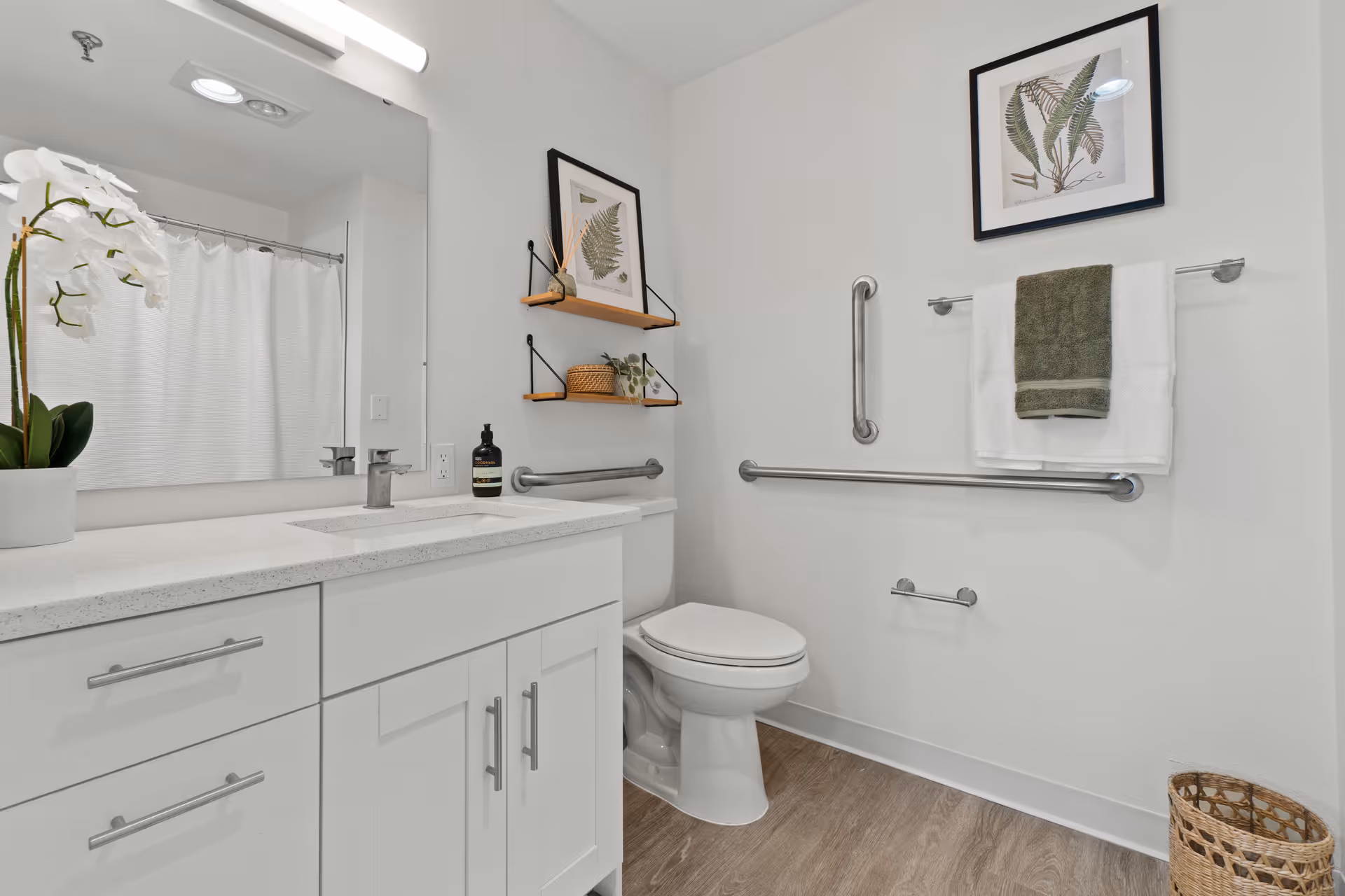 A clean and modern bathroom with white walls and cabinetry, a large mirror above the sink, a white toilet, and safety grab bars installed on the walls. There are two framed botanical prints on the walls, a white orchid plant on the countertop, and neatly folded towels hanging on a towel rack. The floor has a wood-like finish and a wicker basket is placed in the corner.