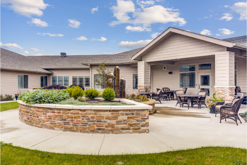 Outdoor patio area at Lakewood Senior Living featuring a stone planter with various plants and a water fountain. The patio has several wicker chairs and tables with cushions under a covered porch attached to a beige building. The sky is partly cloudy.