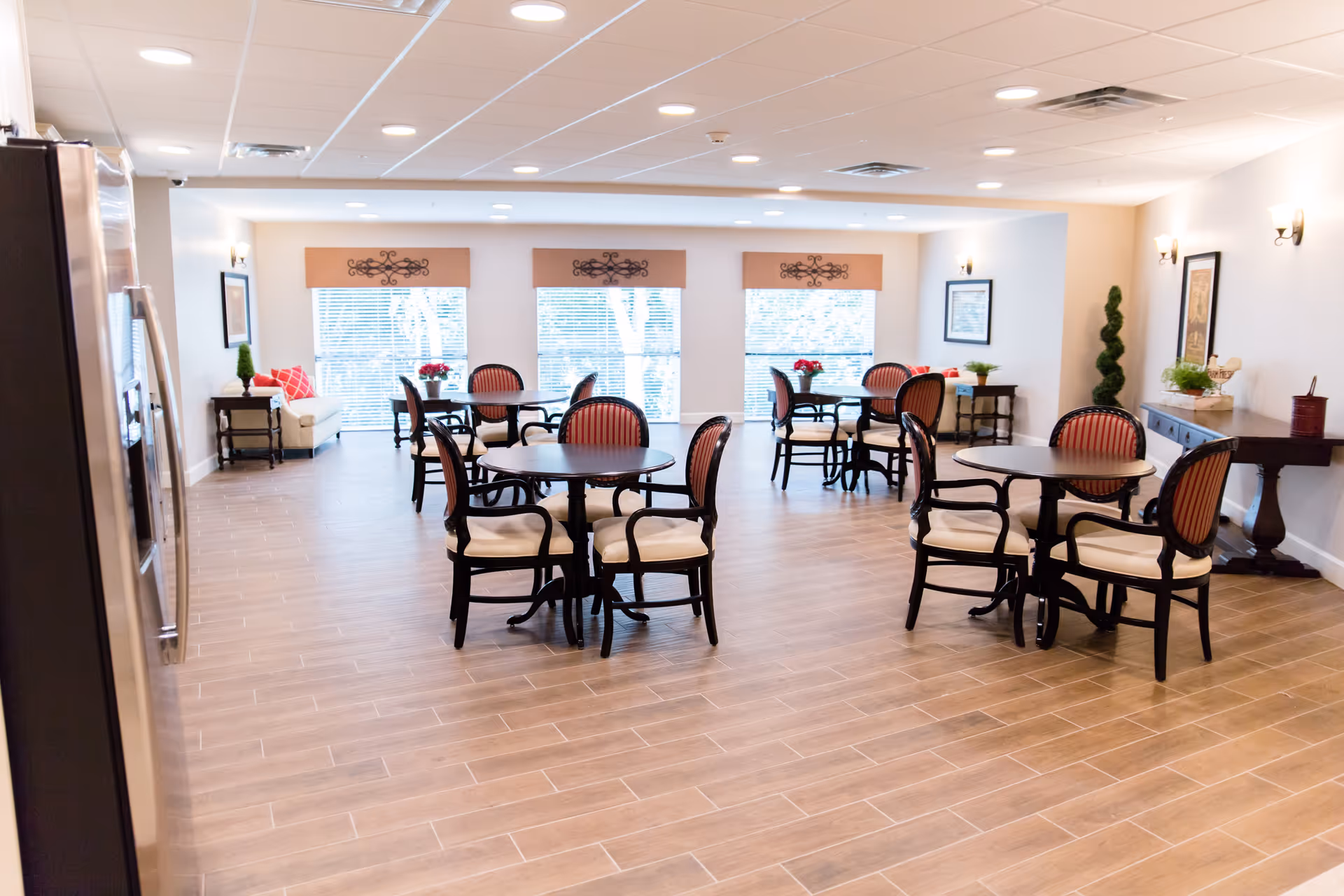 A bright and spacious dining room with several round tables and chairs arranged neatly. The room features large windows with blinds, decorative wall art, and plants on side tables. The floor is covered with light wood-like tiles, and the walls are painted in a soft beige color.