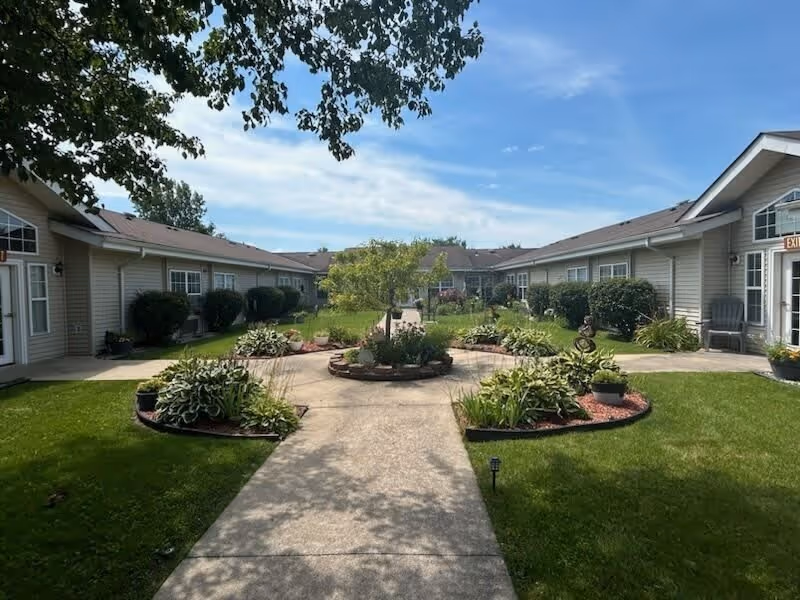 A sunny outdoor courtyard area at Cedar Creek of Marion featuring a concrete walkway leading to a circular garden bed with plants and a small tree in the center. The courtyard is surrounded by single-story beige buildings with windows and doors, green grass, and additional landscaping with bushes and plants.