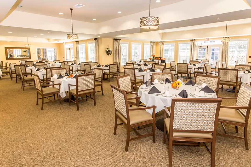 A spacious, well-lit dining room with multiple round tables covered in white tablecloths, each set with black napkins, glassware, and white cups. The room features beige carpet, cushioned wooden chairs with patterned backs, large windows with curtains, and decorative hanging light fixtures.