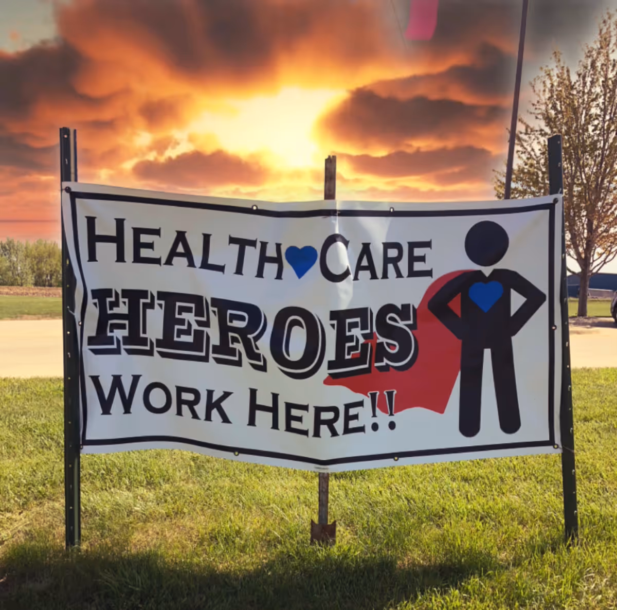 A large outdoor banner on a grassy area with a sunset sky in the background. The banner reads 'HEALTH CARE HEROES WORK HERE!!' with a graphic of a person wearing a red cape and a blue heart on their chest.