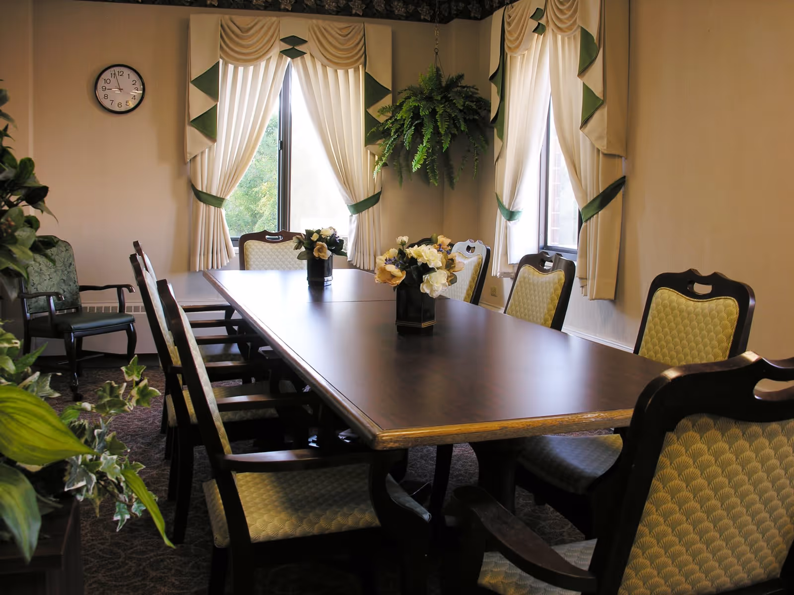 A formal dining room with a long wooden table surrounded by upholstered chairs. The room has two windows with cream-colored curtains accented with green trim. There are floral arrangements on the table and a hanging fern plant in the corner. A clock is mounted on the wall.