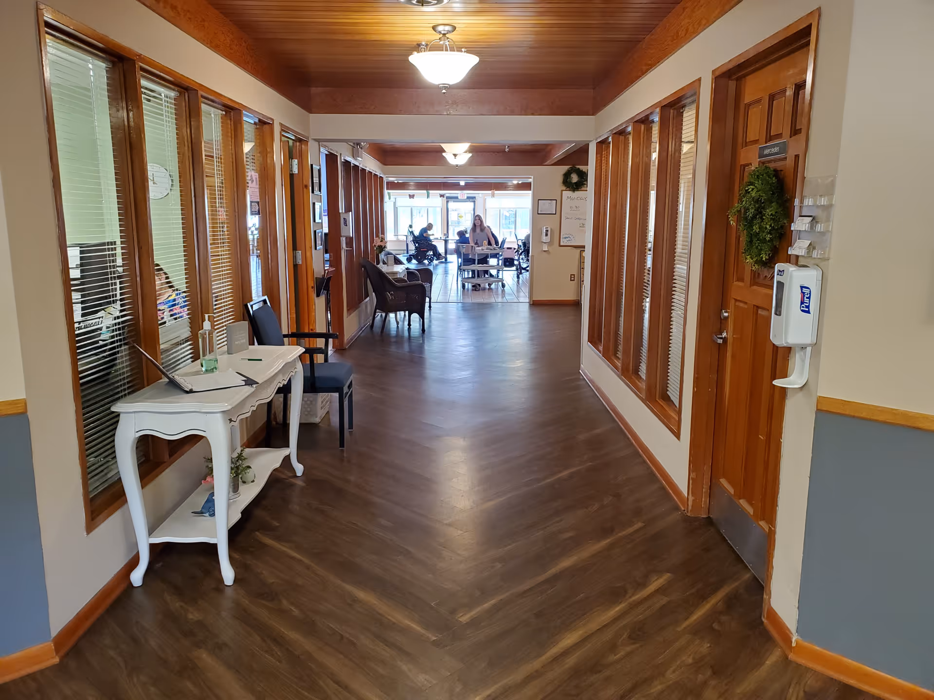 Interior hallway of a senior living facility with wooden floors and ceiling. On the left side, there are glass windows with blinds and a small white table with a laptop, hand sanitizer, and a chair. On the right side, there is a wooden door with a green wreath and a mounted Purell hand sanitizer dispenser. At the end of the hallway, there is a common area with people sitting and interacting.