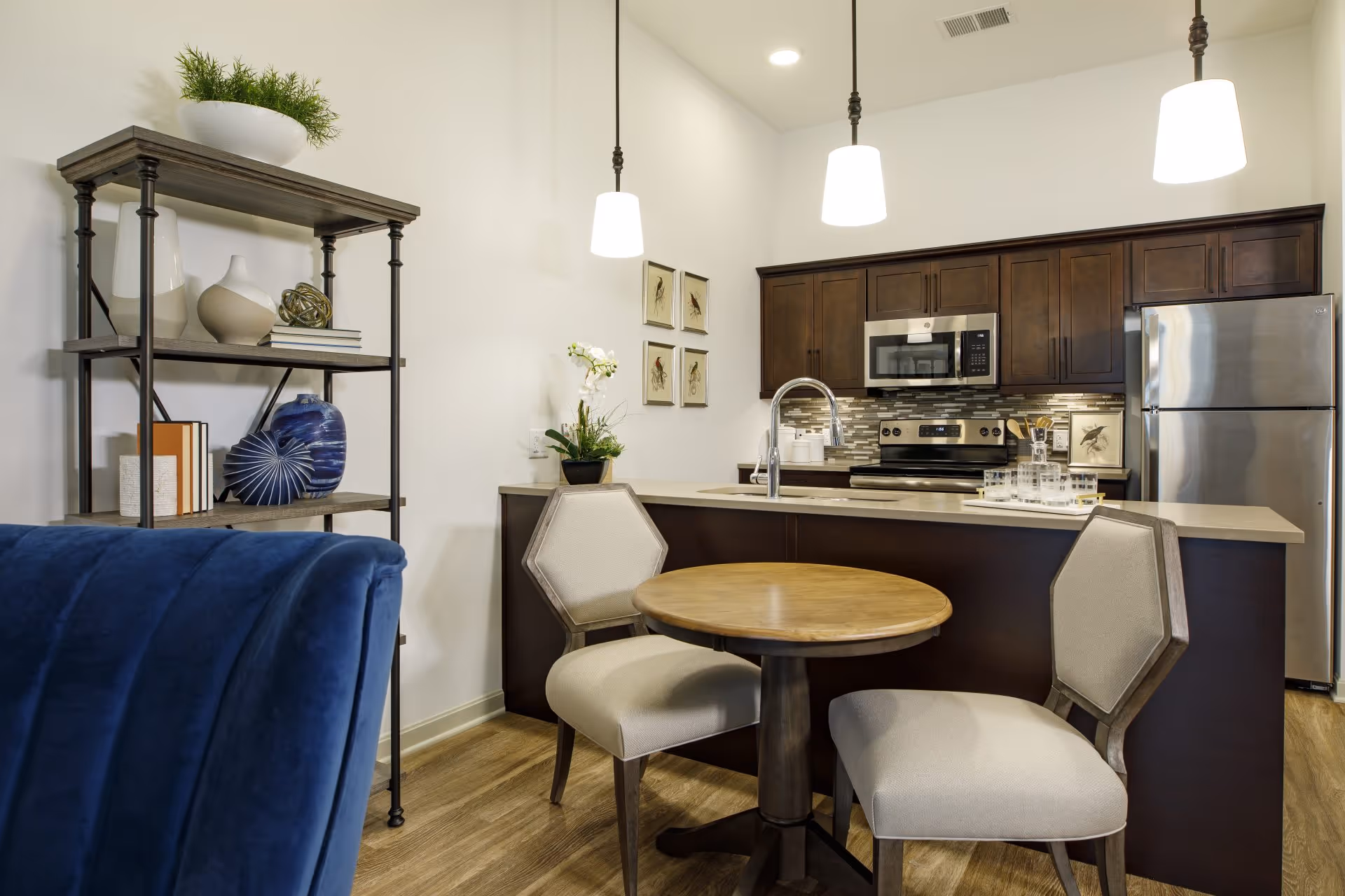 A modern kitchen and dining area in an assisted living facility featuring a small round wooden table with two upholstered chairs, a kitchen island with a sink, dark wood cabinets, stainless steel appliances including a refrigerator and microwave, three pendant lights hanging from the ceiling, and a decorative shelving unit with vases and books.