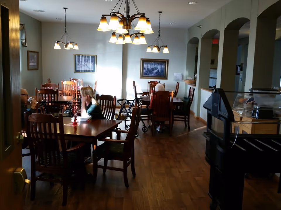 Interior view of a dining room in a senior living facility with wooden tables and chairs. Several elderly people are seated at the tables, some using walkers. The room is lit by hanging light fixtures and has framed pictures on the walls.