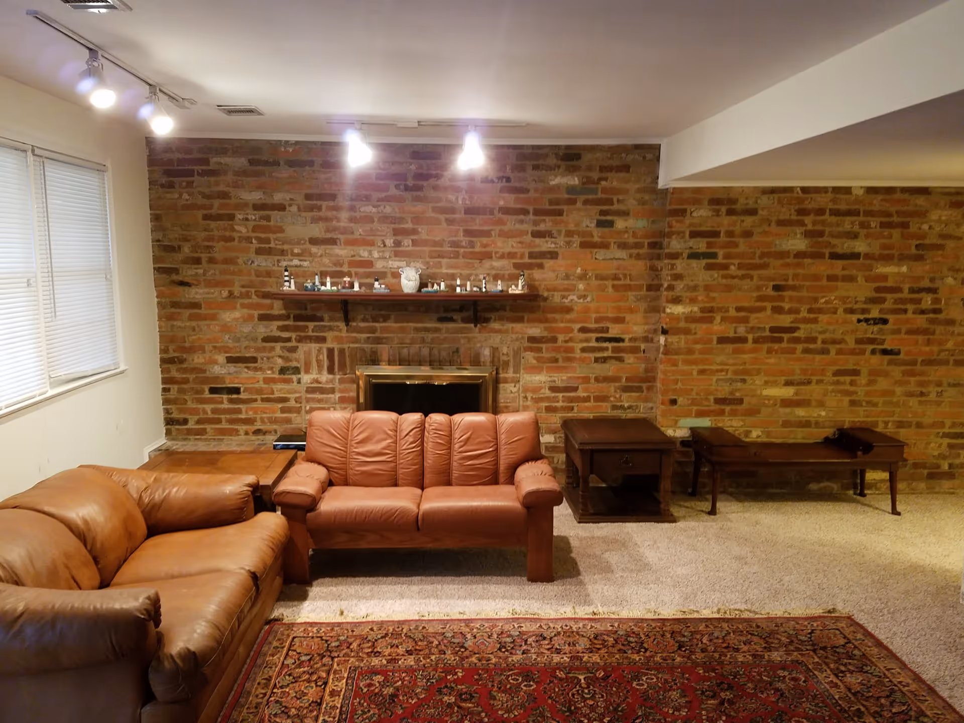 A cozy living room with a brick wall featuring a fireplace in the center. Above the fireplace is a wooden shelf displaying small decorative items. In front of the fireplace are two brown leather sofas, one larger and one smaller. To the right of the smaller sofa are two wooden side tables. The floor is carpeted, and a patterned area rug is partially visible in the foreground. There is a window with closed blinds on the left wall and ceiling lights illuminating the room.