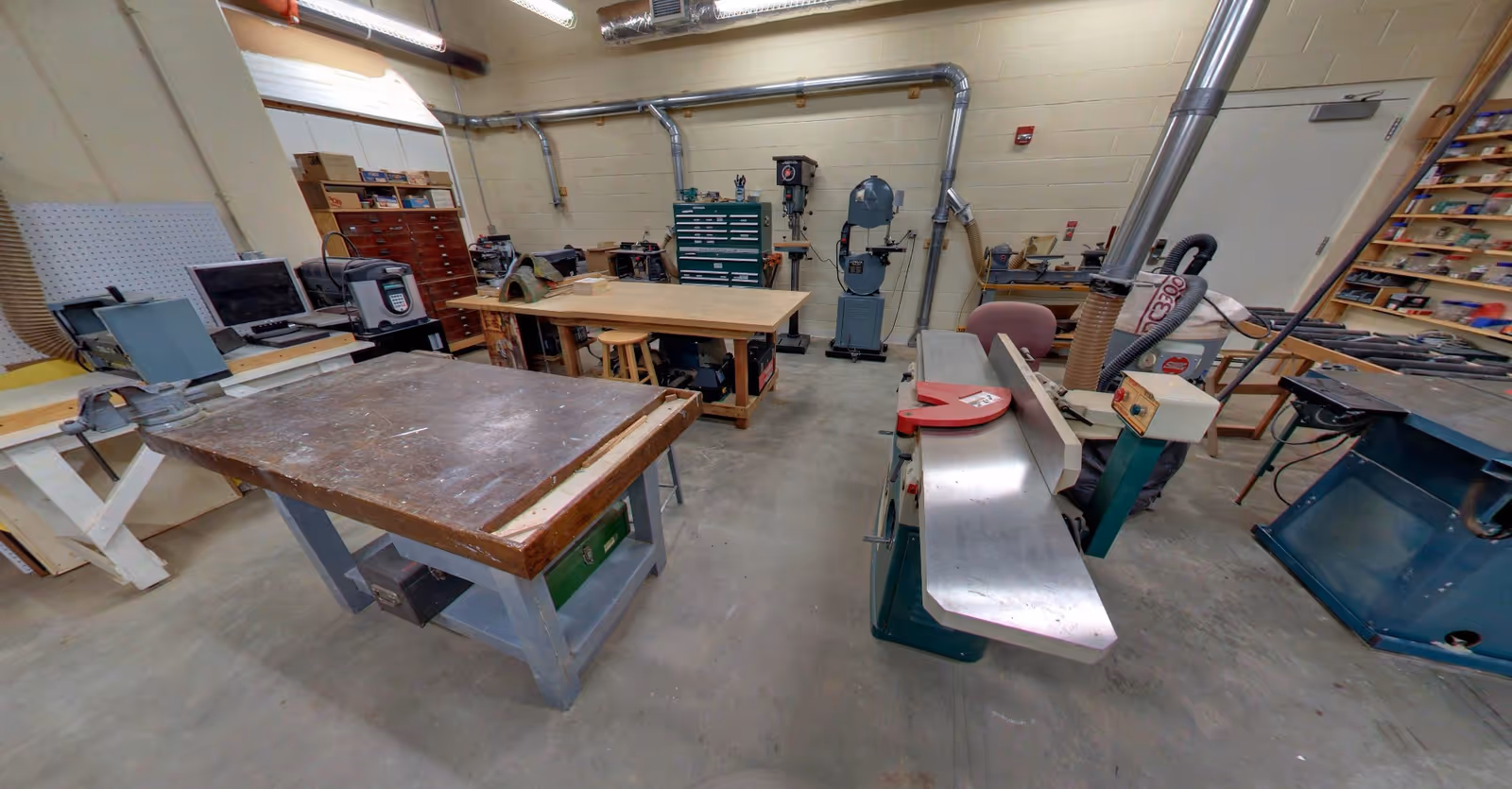 Interior view of a workshop room with various woodworking tools and equipment including workbenches, a planer, a band saw, and shelves with tools and supplies. The room has concrete floors, beige walls, and exposed ductwork on the ceiling.