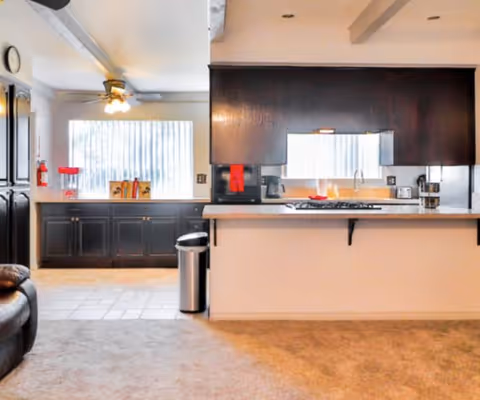 Interior view of a senior living facility kitchen area with dark wood cabinets, a countertop with a built-in stove, a coffee maker, toaster, and a trash can. There is a window with vertical blinds letting in natural light and a ceiling fan with lights above the tiled floor section.