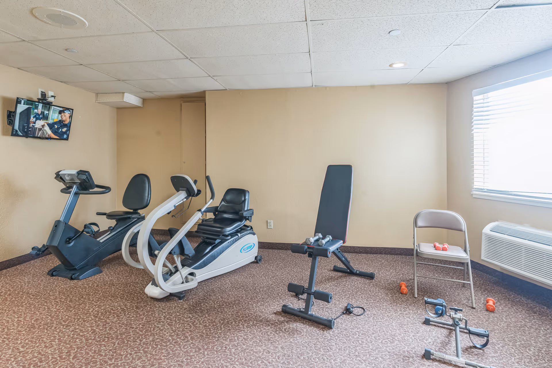 A small exercise room with two stationary recumbent bikes, a weight bench with dumbbells, a folding chair with small dumbbells on the seat, and a pedal exerciser on the carpeted floor. A wall-mounted TV is on the left wall, and a window with blinds is on the right wall.