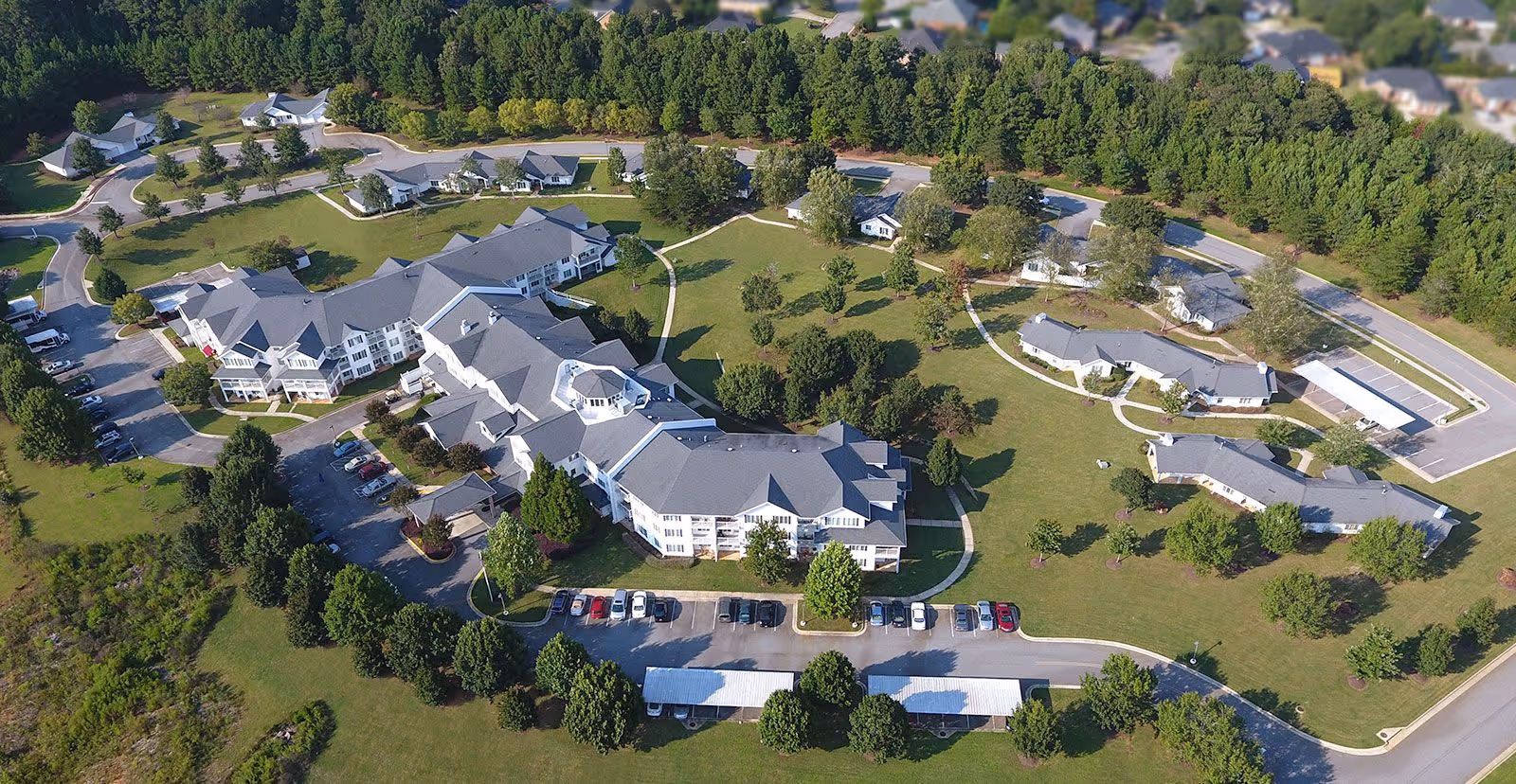 Aerial view of TerraBella Epps Bridge senior living facility showing multiple buildings with gray roofs surrounded by green lawns, trees, and parking areas. The complex is bordered by a dense line of trees and has curved roads and walkways connecting the buildings.