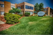 Exterior view of Allison Care Center building with a well-maintained lawn, bushes, and trees. The building is two stories with large windows and a sign in front displaying the facility's name.