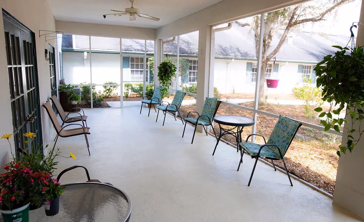 A screened-in porch area with several chairs and small tables arranged along the walls. There are hanging plants and potted flowers, with a ceiling fan overhead. Outside the screened porch, there is a view of a garden area and a building with windows and blue shutters.