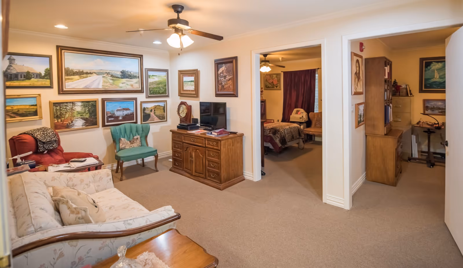 Cozy communal living area with a sofa, chairs, framed wall art, a TV cabinet, and open doorways to adjacent rooms.
