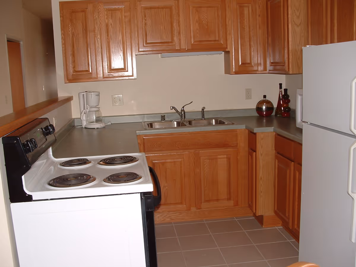 A kitchen with wooden cabinets, a white electric stove with four coil burners, a double stainless steel sink, a white refrigerator, a coffee maker on the counter, and decorative bottles on the countertop.