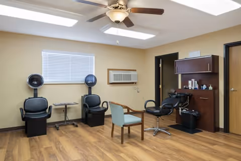 Small salon-style room with hooded hair dryers, styling chairs, a shampoo sink and cabinetry on wood flooring.