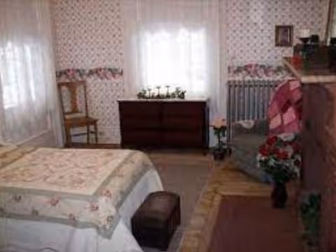 A cozy bedroom with floral wallpaper and a bed covered with a quilted bedspread. There is a wooden chair near a window with sheer white curtains, a dark wooden dresser beneath another window, and a cushioned armchair with a pink and white quilt draped over it. The room also features a small footstool, a radiator, and some potted plants on the floor.