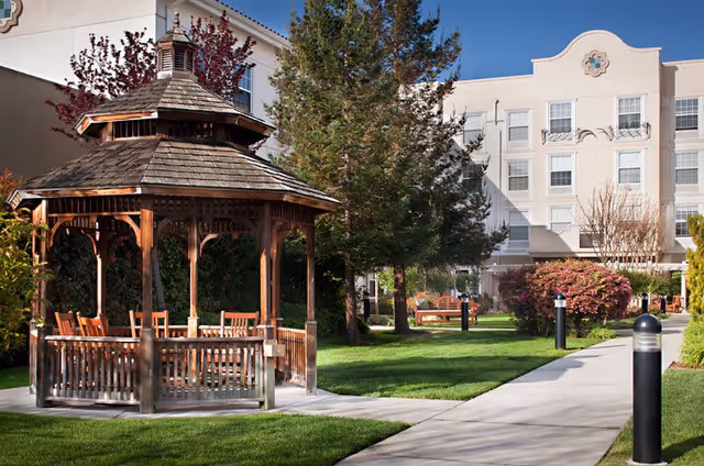 Outdoor garden area at Belmont Village Senior Living Sunnyvale featuring a wooden gazebo with chairs inside, surrounded by green grass, trees, bushes, and a paved walkway leading to a multi-story building in the background under a clear blue sky.