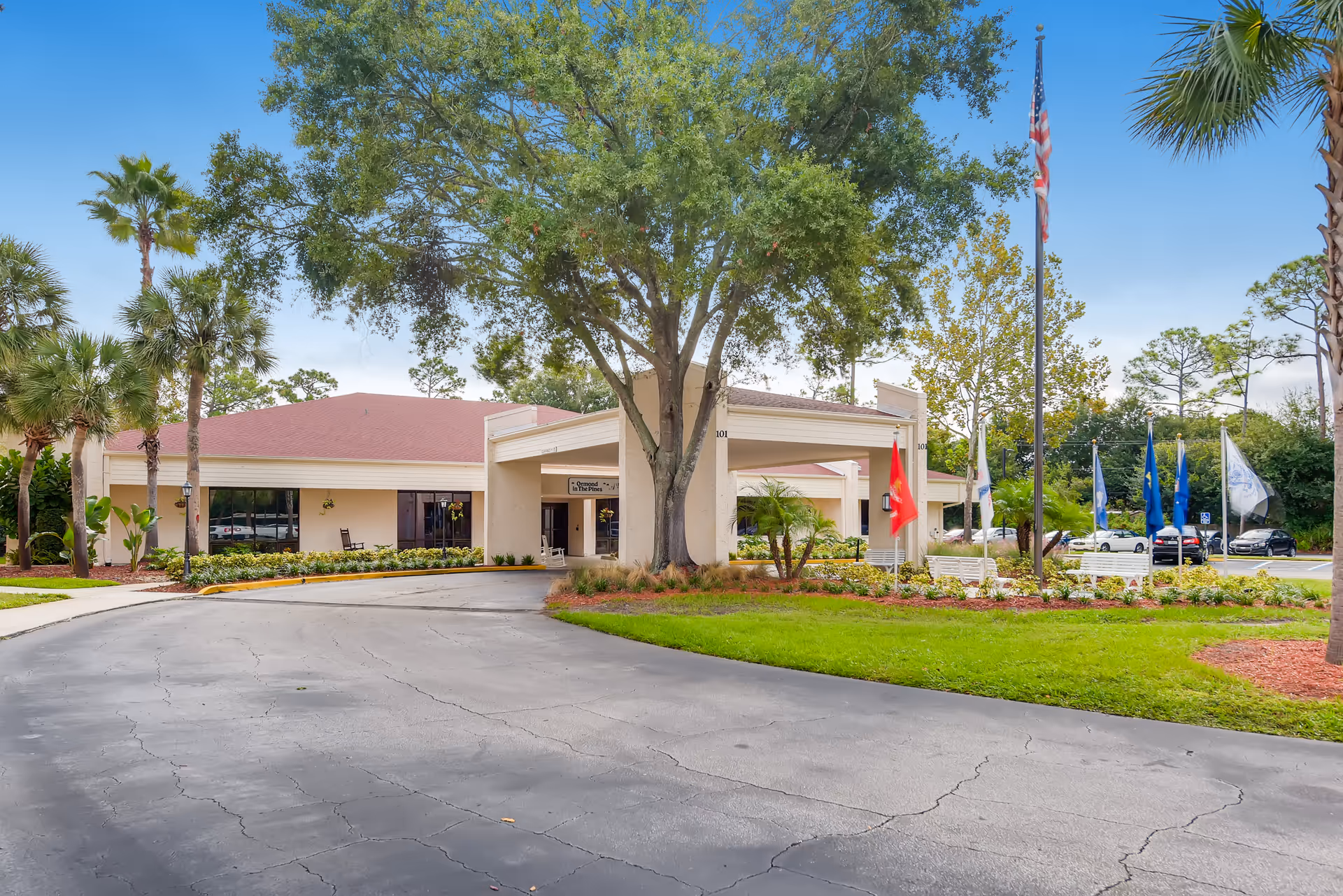 Exterior view of Ormond In The Pines Senior Living facility showing a single-story building with a covered entrance, surrounded by trees, palm trees, and a well-maintained lawn. Several flags are displayed on flagpoles near the entrance, and a paved driveway leads up to the building.