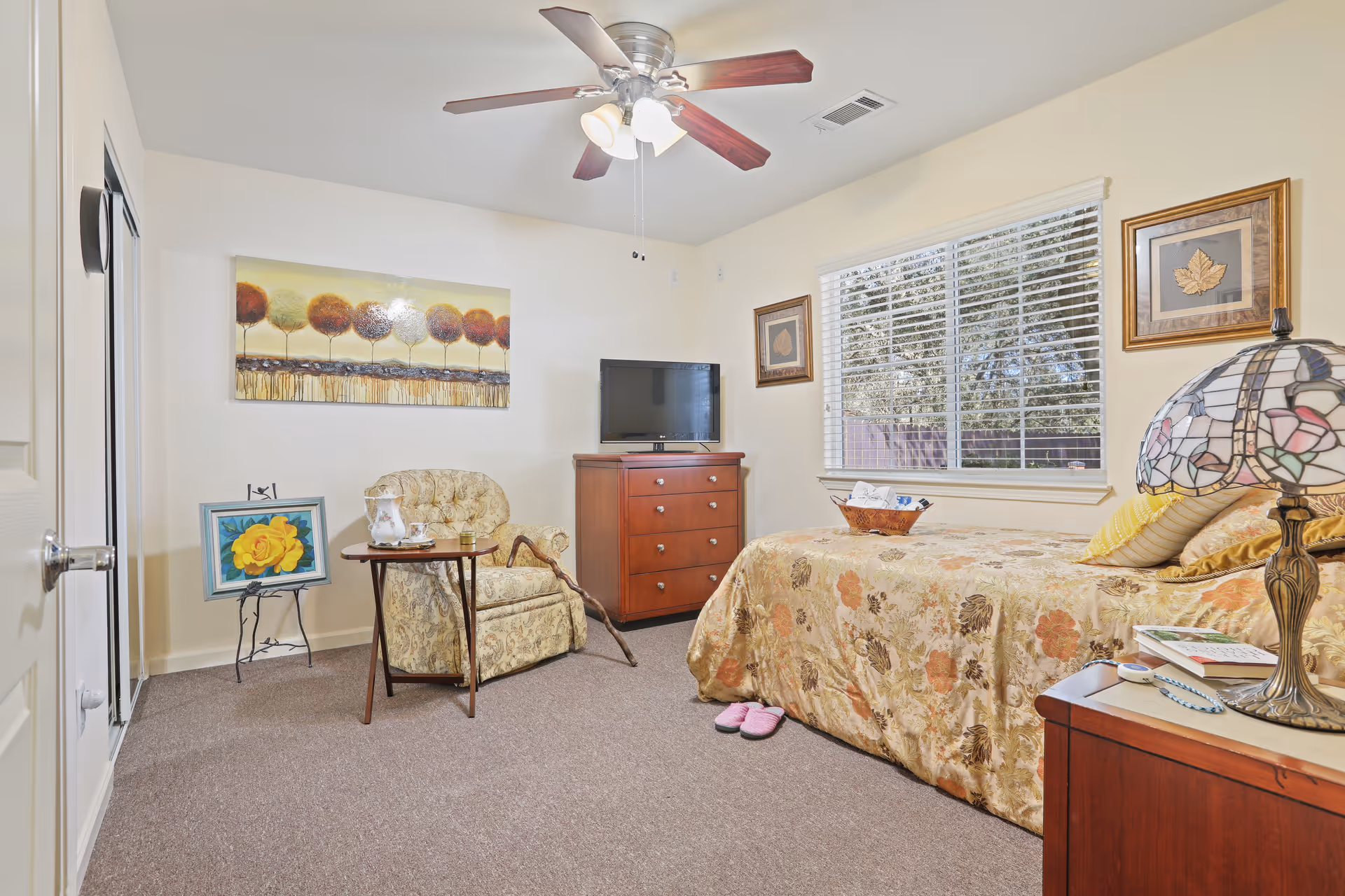 A cozy bedroom in Citrus Pines Senior Living featuring a single bed with floral bedding, a wooden nightstand with a decorative lamp and books, a wooden dresser with a flat-screen TV, a comfortable armchair with a small side table holding a pitcher and glass, and artwork on the walls. A ceiling fan with lights is mounted above, and a window with blinds lets in natural light.