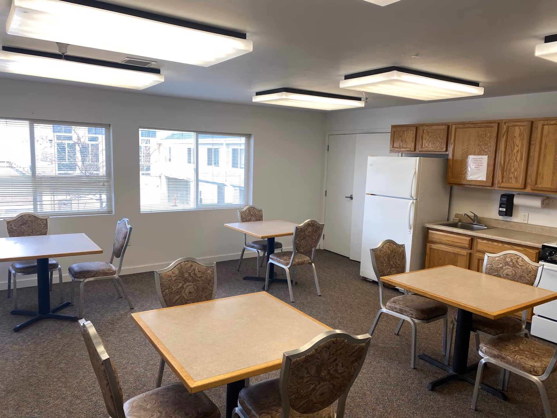 Communal dining room with several small square tables and upholstered chairs, a kitchenette with refrigerator and cabinets, and large windows letting in daylight.