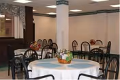 A dining room with multiple round tables covered with white tablecloths and blue placemats. Each table has a small floral centerpiece. The room has beige walls, a window with patterned curtains, and several black chairs around the tables.