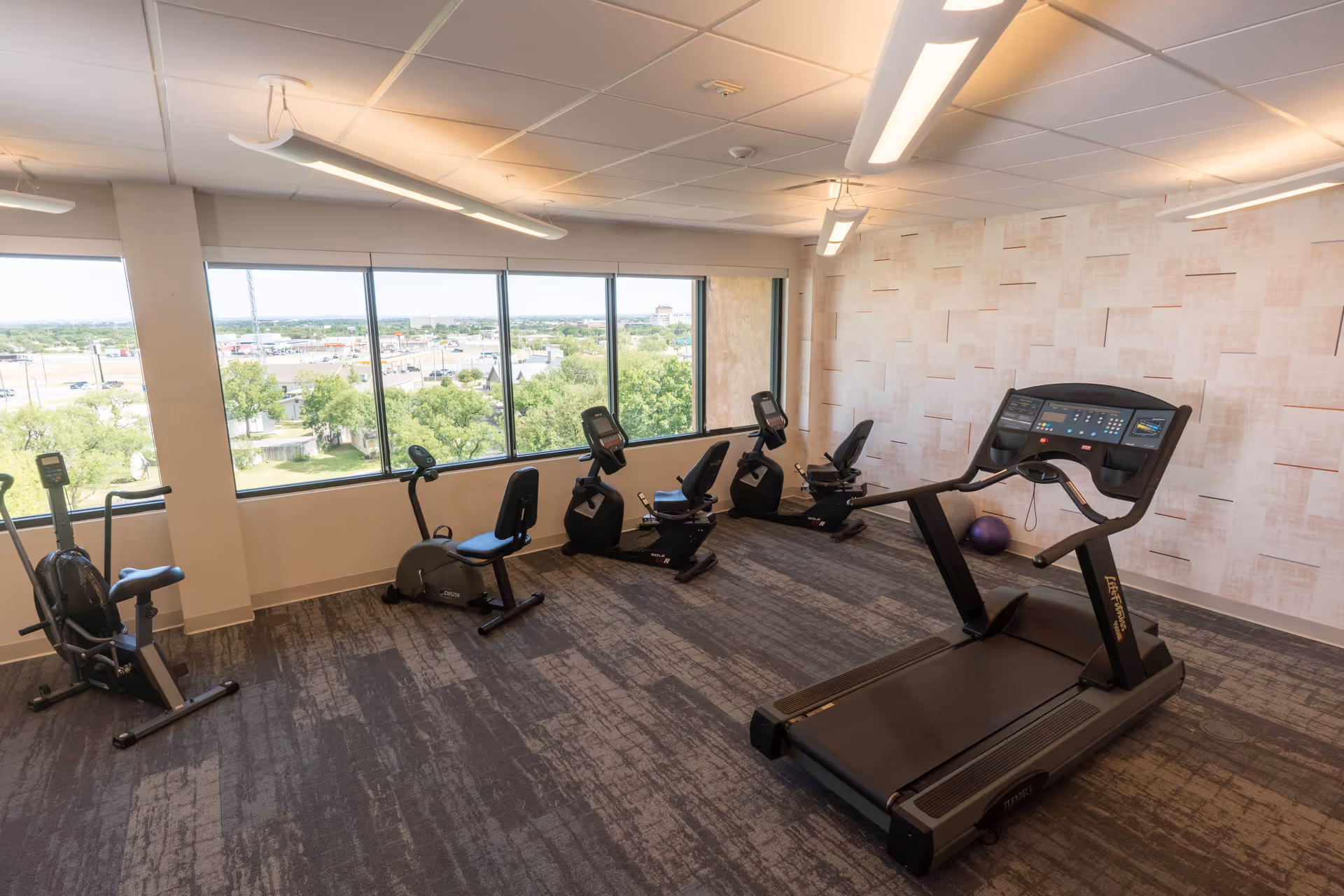 A fitness room with exercise equipment including a treadmill and several stationary bikes. The room has large windows providing a view of trees and buildings outside. The walls are light-colored with a textured pattern, and the ceiling has fluorescent lighting.