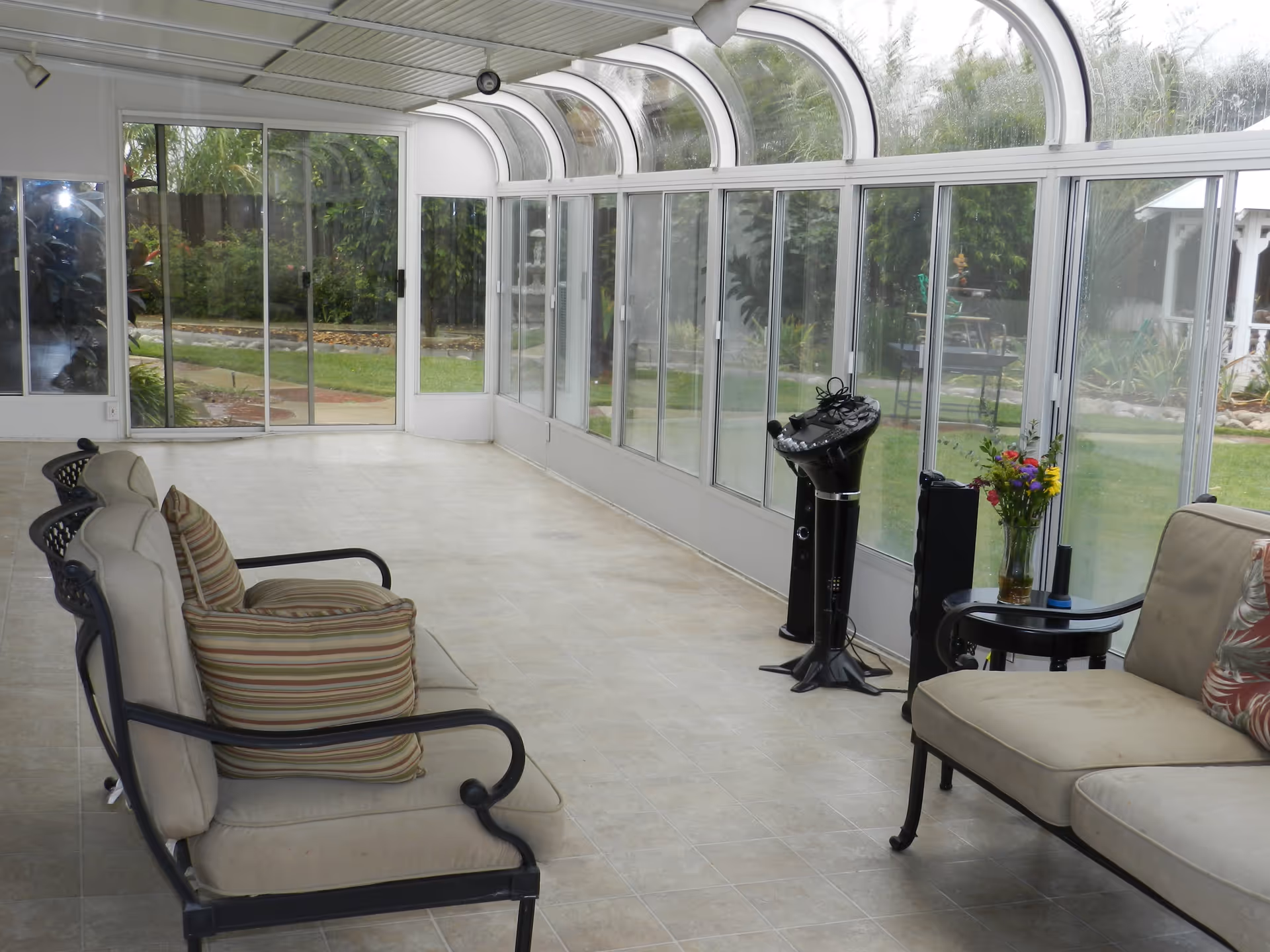 A sunroom with large curved glass windows and sliding glass doors overlooking a garden. The room has tiled flooring and contains cushioned metal-framed chairs with striped pillows, a small round table with a vase of flowers, and a black standing karaoke machine or speaker.