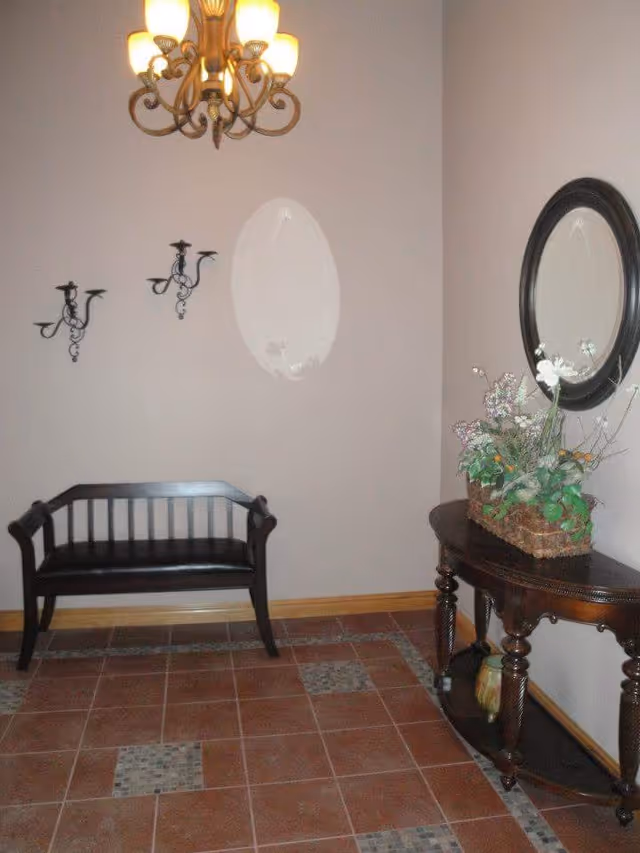 An entryway or small sitting area with a dark wooden bench against a beige wall, a decorative chandelier hanging from the ceiling, two black wall-mounted candle holders, and a wooden console table with a floral arrangement and a round mirror above it. The floor is tiled with a pattern of reddish-brown and gray tiles.