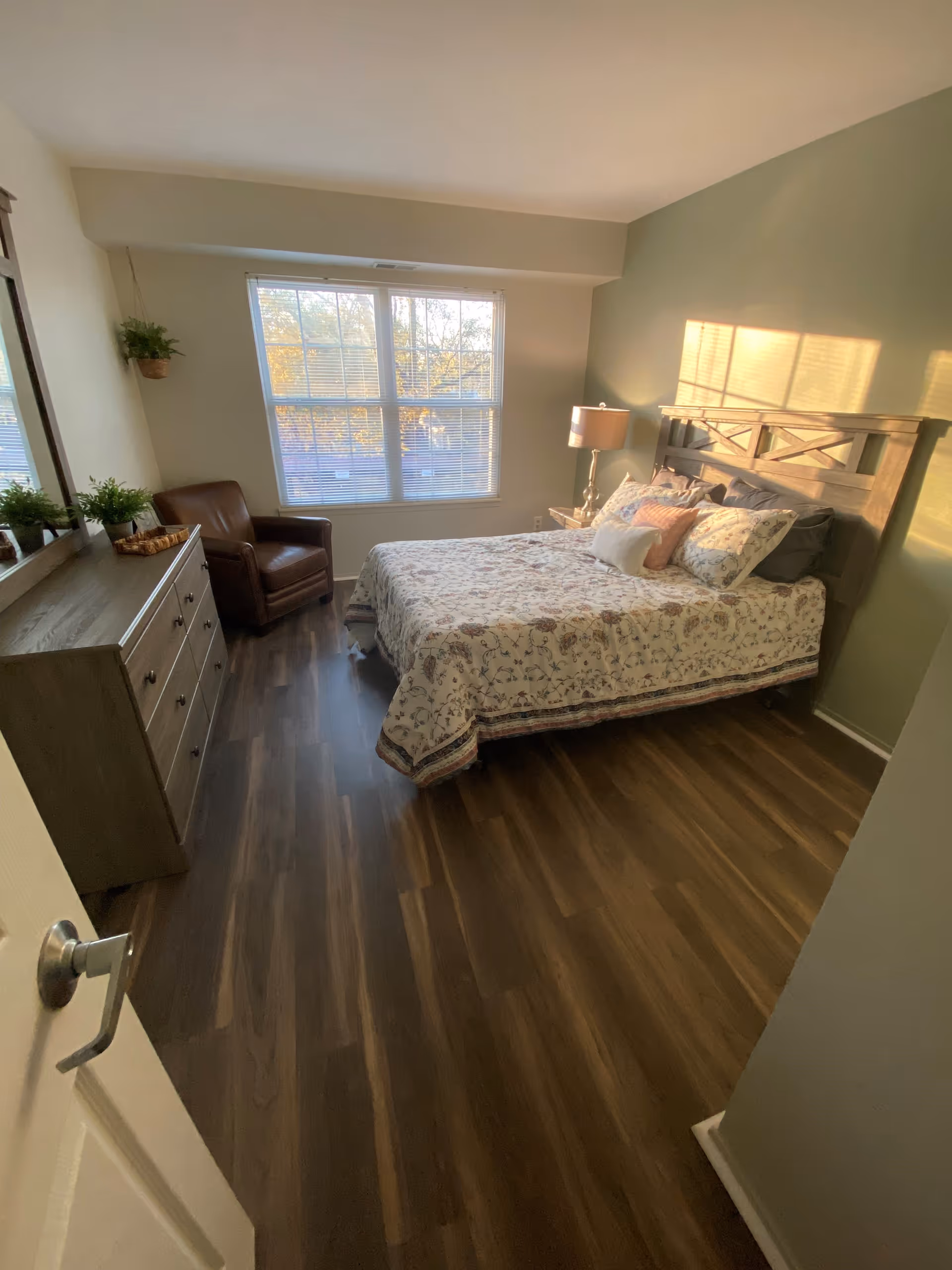 A cozy bedroom with a large window letting in natural light. The room features a wooden bed with a floral bedspread and multiple pillows, a wooden dresser with plants on top, a brown leather armchair, and a bedside table with a lamp. The floor is dark wood, and the walls are painted in light colors.