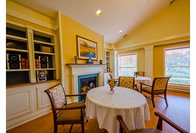 A cozy room with yellow walls featuring a white fireplace with a landscape painting above it. There are two round tables covered with white tablecloths, each with a small flower arrangement. Four wooden chairs with patterned cushions surround the tables. Large windows with blinds let in natural light, and built-in shelves with decorative items are visible.