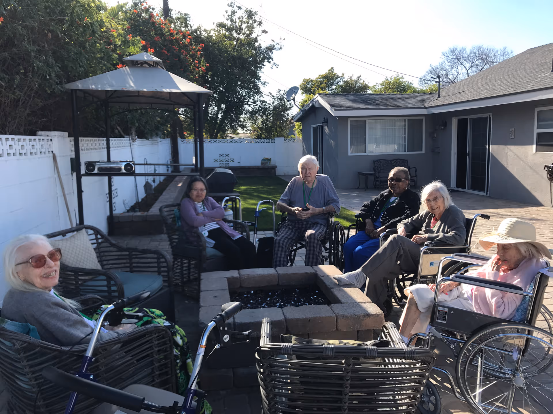 A group of six elderly individuals sitting outdoors around a square fire pit. Some are in wheelchairs and others are seated on outdoor chairs. They are in a patio area with a gazebo, a white fence, and a gray building in the background. The weather is sunny and clear.
