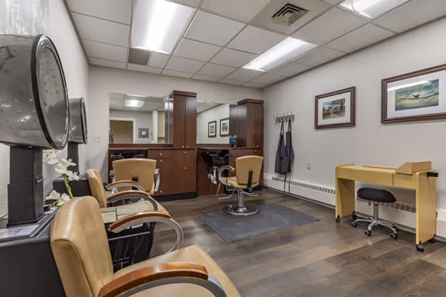 Interior view of a salon area in a senior living facility with salon chairs, hair dryers, a manicure station, and framed artwork on the walls.