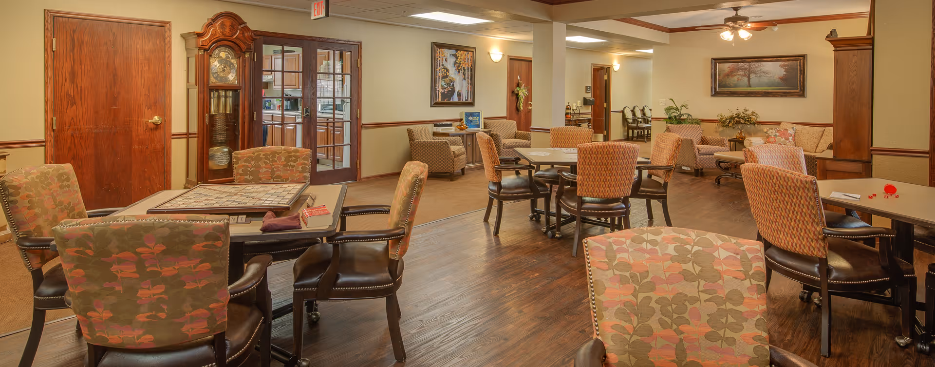 A cozy common area in a senior living facility featuring multiple tables and chairs with patterned upholstery. There is a large wooden grandfather clock near a set of glass double doors leading to a kitchen. The room has warm lighting, framed artwork on the walls, and a ceiling fan with lights. In the background, there are additional seating areas with armchairs and a sofa, along with plants and decorative items.
