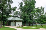 A small pavilion with a green metal roof sits by a curved walkway amid trees next to a pond.