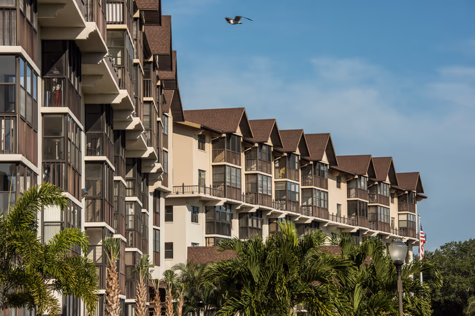 Exterior view of a multi-story residential building with balconies and brown roofs, surrounded by palm trees under a blue sky with a bird flying above.